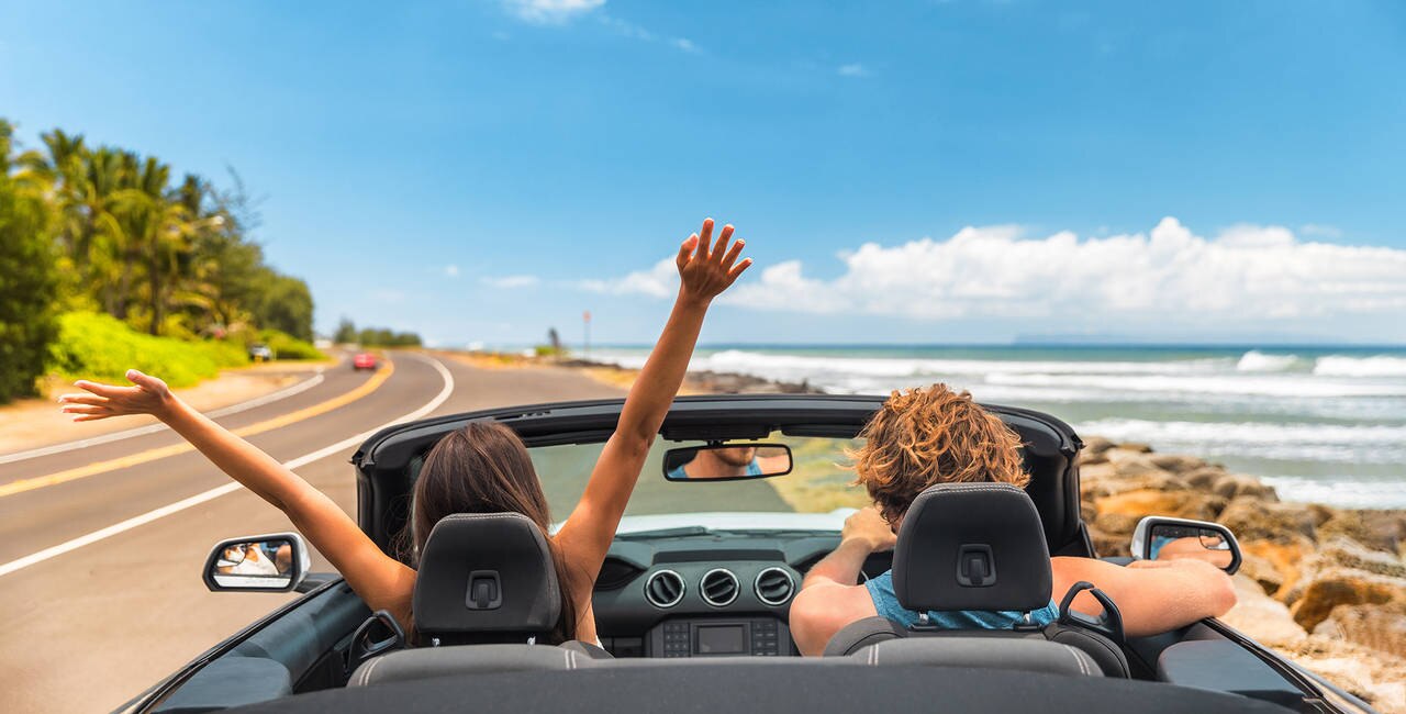 Road trip car holiday happy couple driving convertible car on summer travel Hawaii vacation. Woman with arms up having fun, young man driver