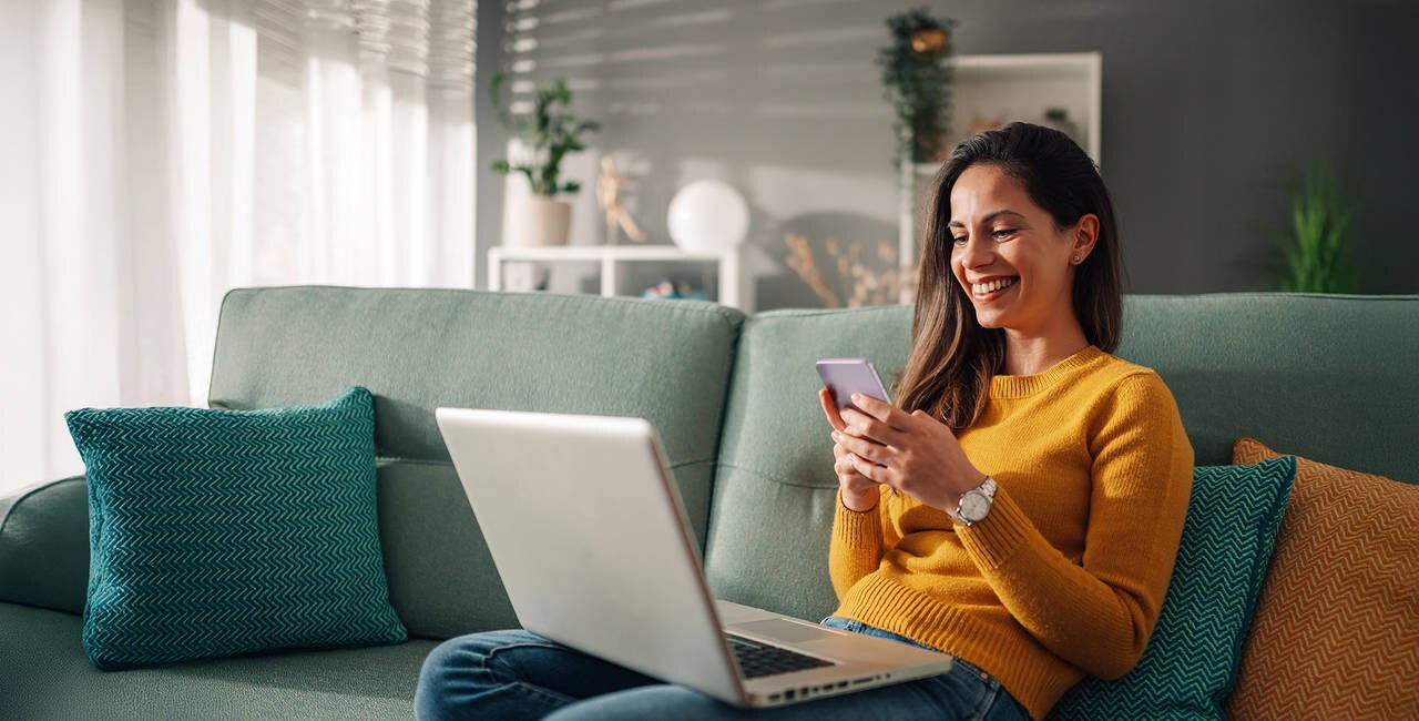 Portrait of a woman using laptop and a phone and checking email or news online while sitting on sofa at home. Searching for friends in internet social networks or working on computer. Copy space.